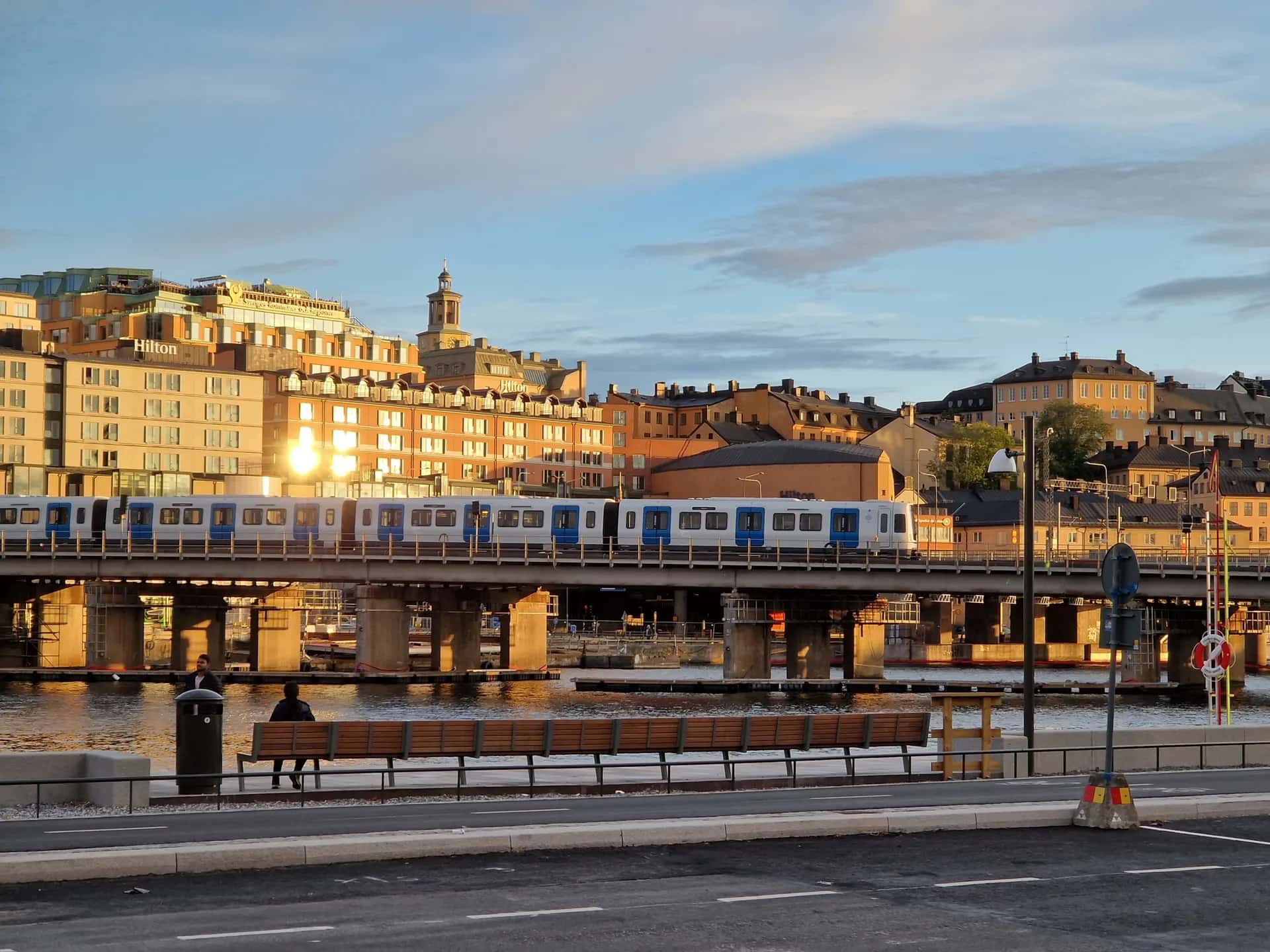 Tunnelbanetåg på bro över vatten med stadssiluett och solglitter i fönster. Två personer vid kajen