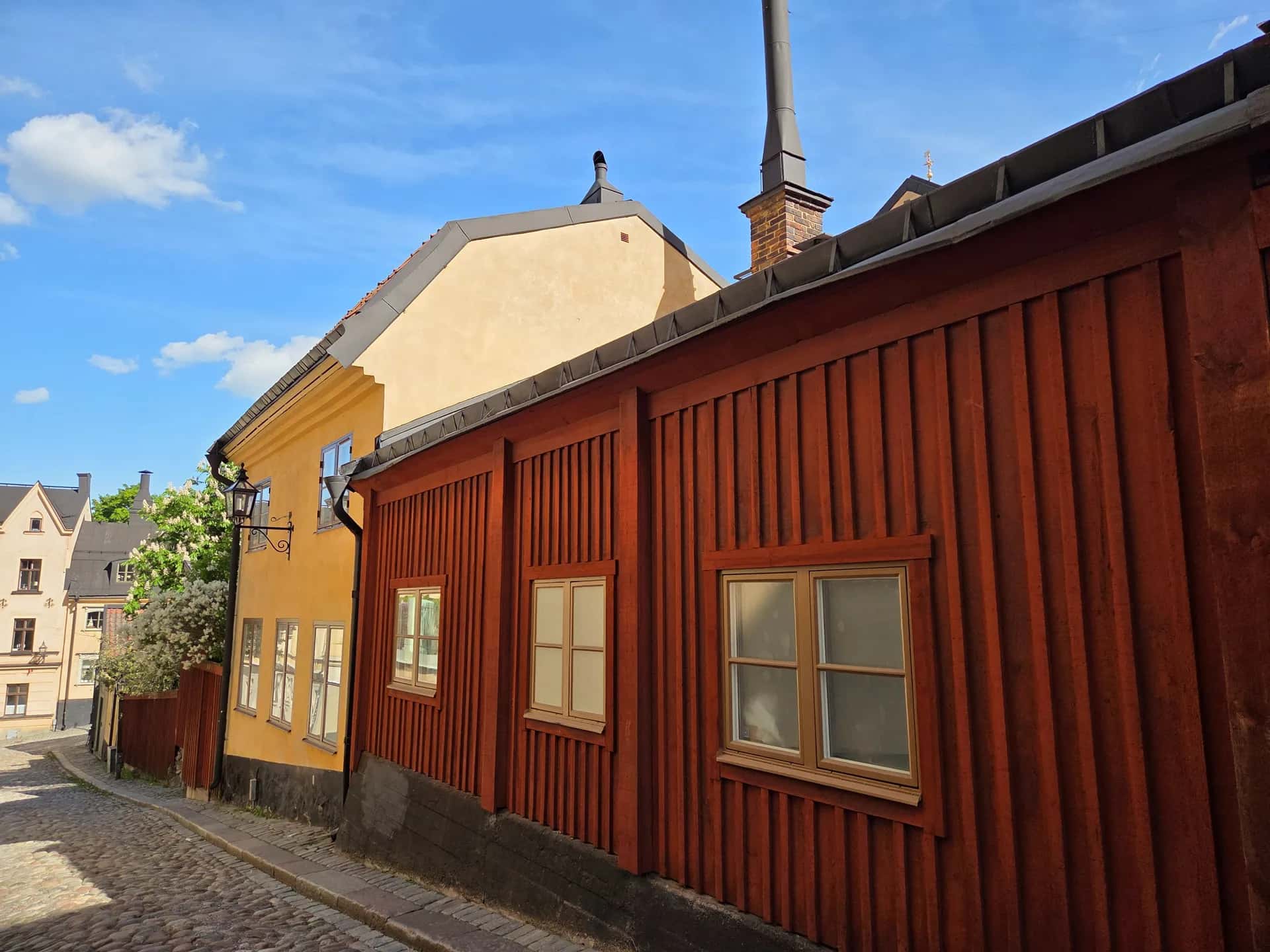 Kullerstensgata med röd träfasad och gul byggnad med fönster under blå himmel med moln