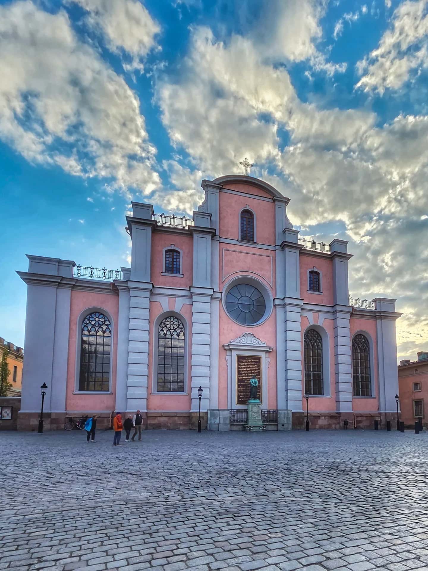 Stor rosa kyrka med gråa detaljer och ett kors på taket, på en kullerstensplats med några personer, under blå himmel med vita moln