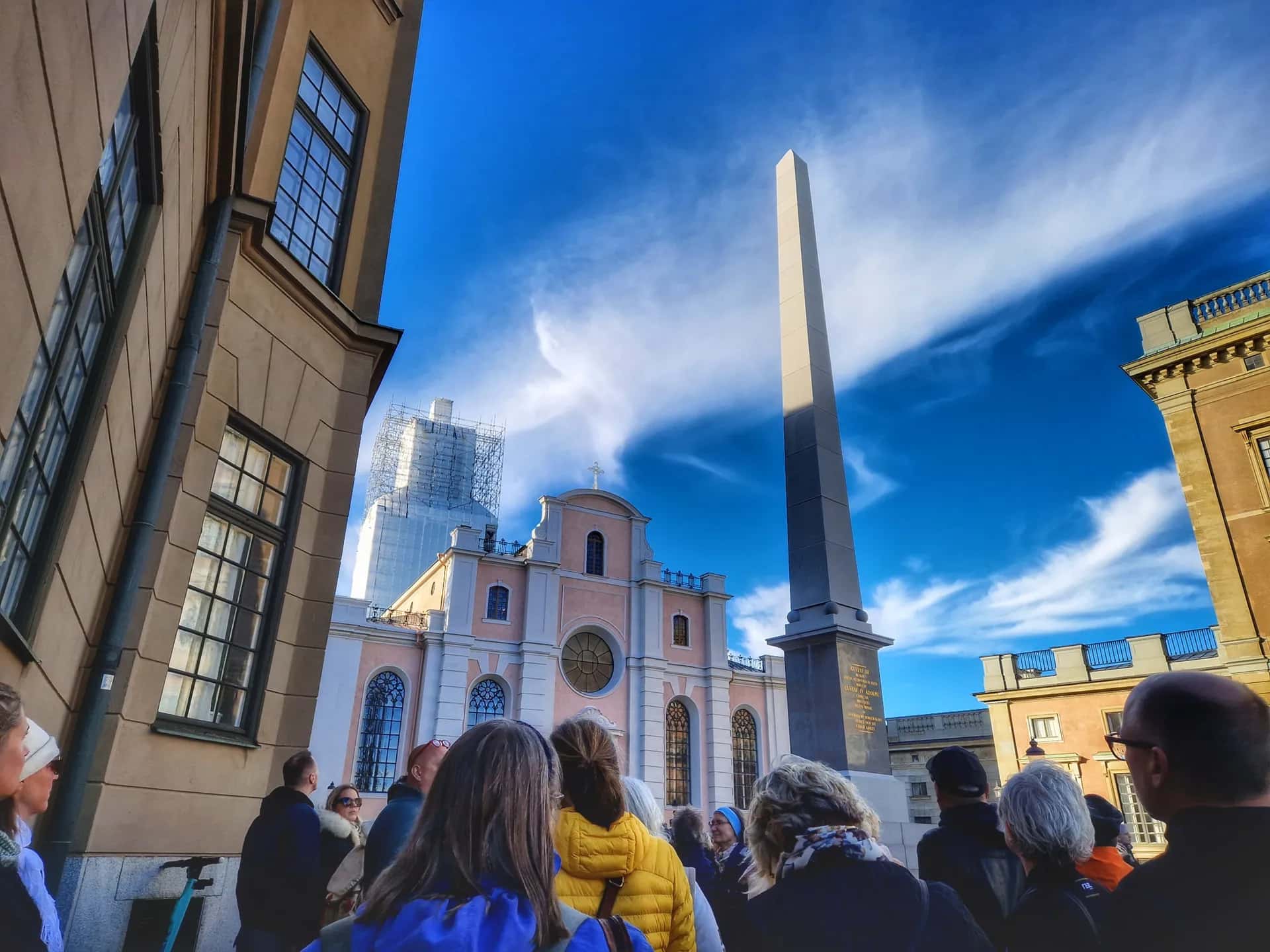 Människor ser mot en obelisk och en rosa byggnad under blå himmel med vita moln. En byggnad har byggnadsställning