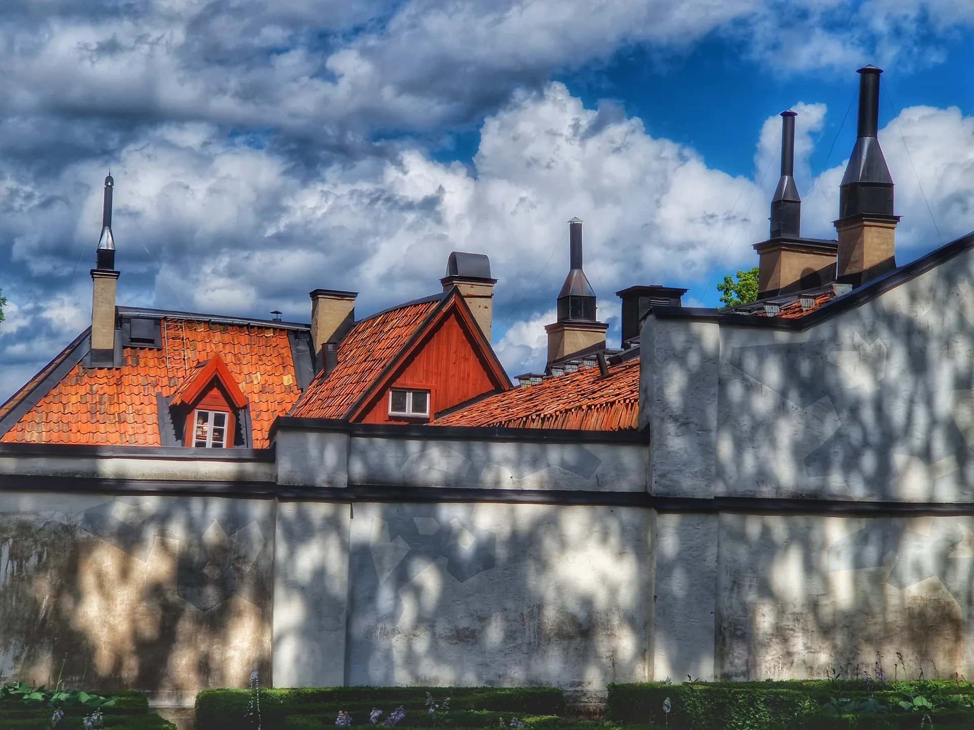 Terracottafärgade tegeltak med skorstenar och vit putsfasad med träskuggor under blå himmel