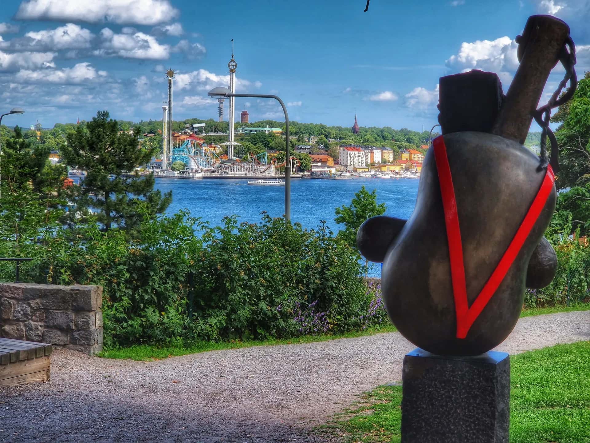 Mörk rundad skulptur med röda V-band på sockel. Bakom den vatten med båtar, nöjesparkens torn och stad