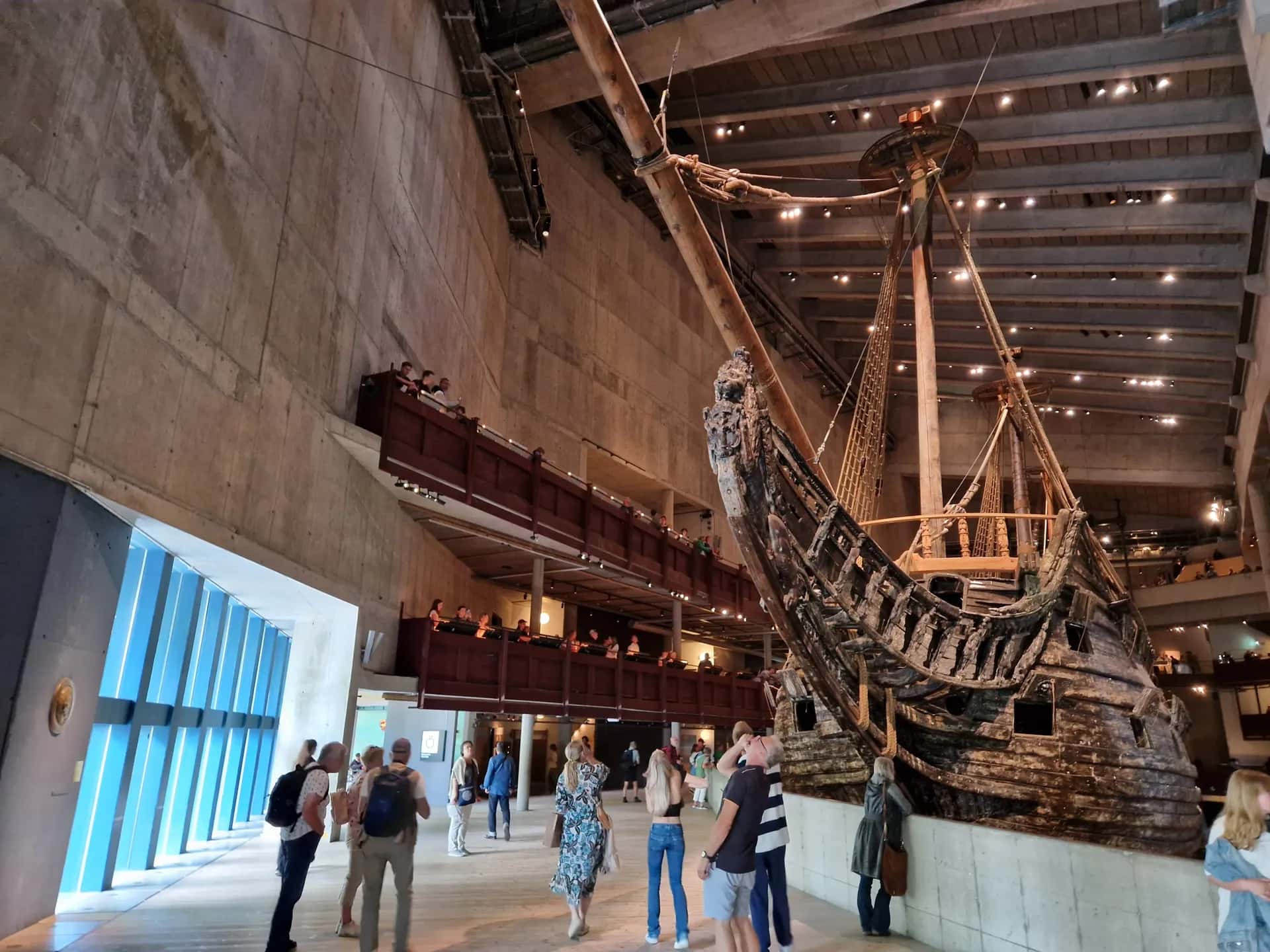 Stort gammalt träskepp i en museihall. Besökare står på golvet och balkonger runt skeppet. Blått fönster till vänster