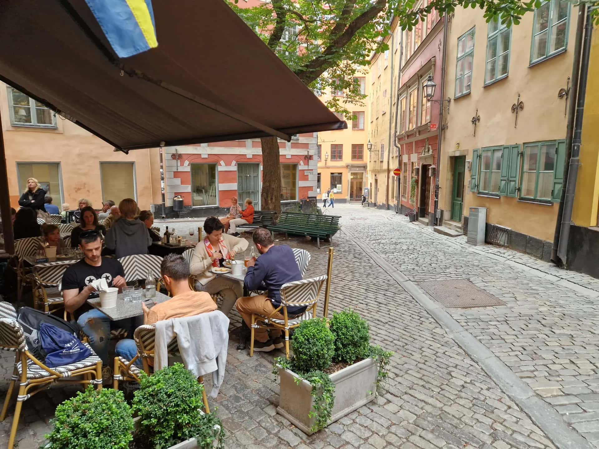 Gäster på uteservering under markis på en kullerstensgata. Blågul flagga, gamla hus och gröna växter syns