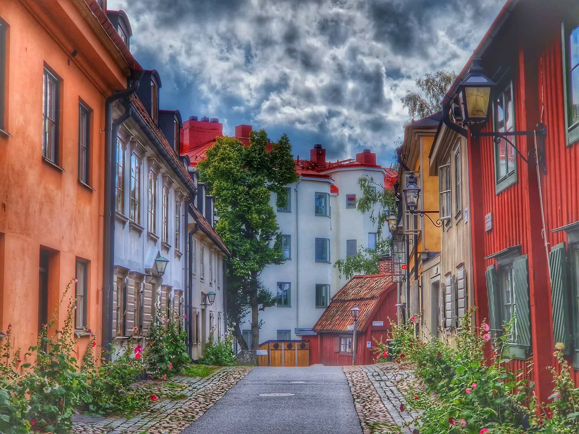 En smal kullerstensgata med färgglada hus, blommor längs väggarna och en molnig himmel