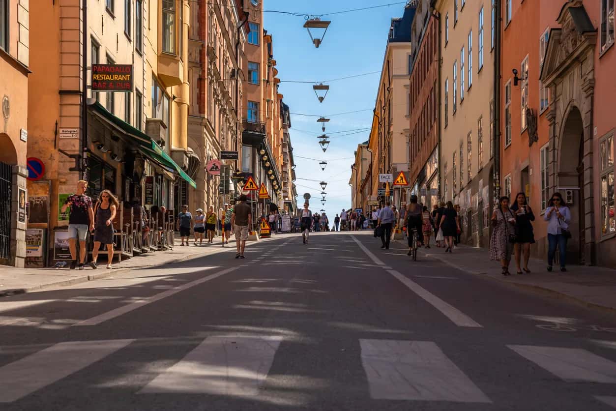 Solig stadsgata med människor, byggnader, ett övergångsställe och dekorativa lampor över vägen