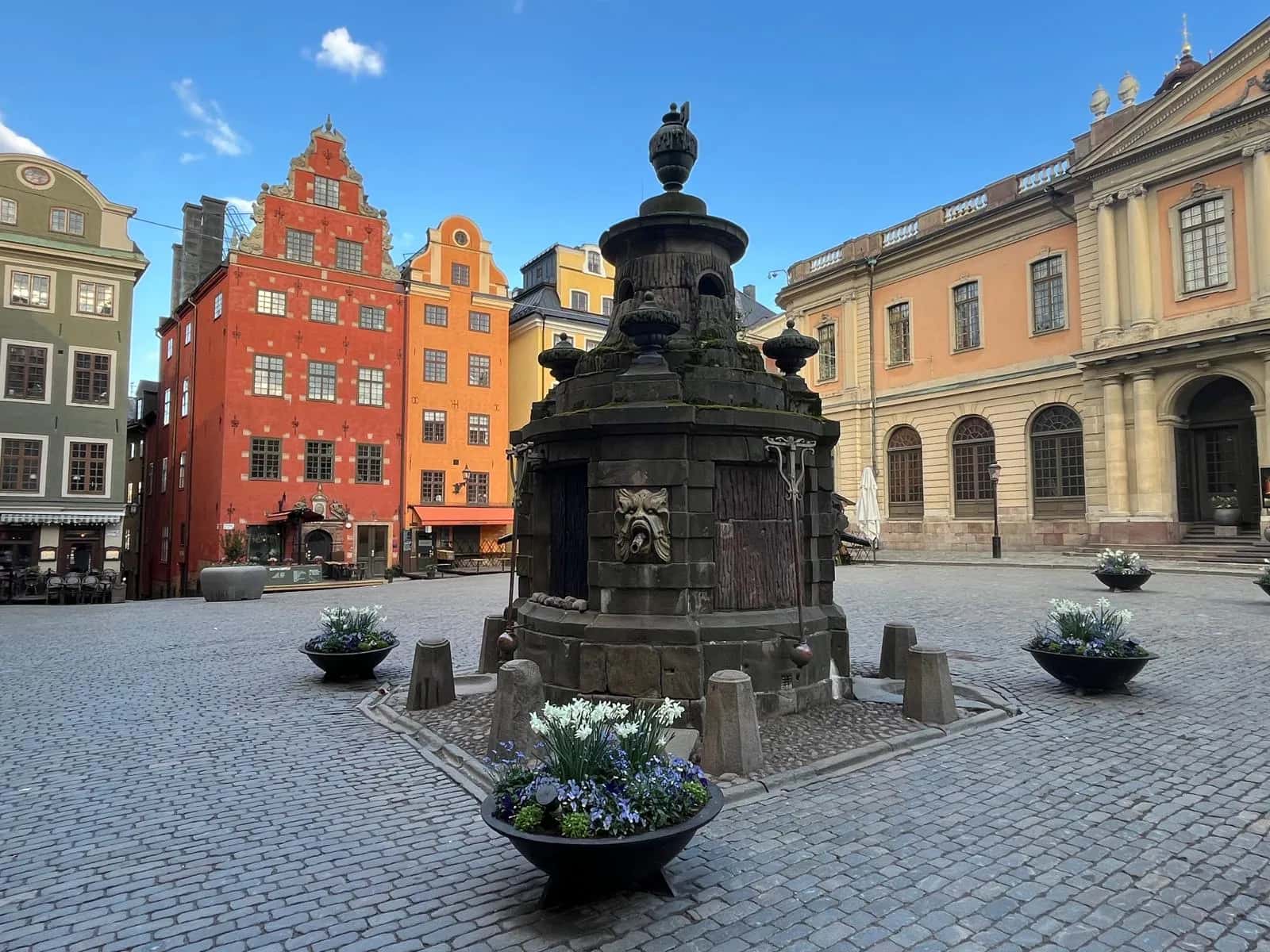 Stor stenfontän på ett kullerstensbelagt torg. Färgglada byggnader och blomkrukor med vita och lila blommor under blå himmel