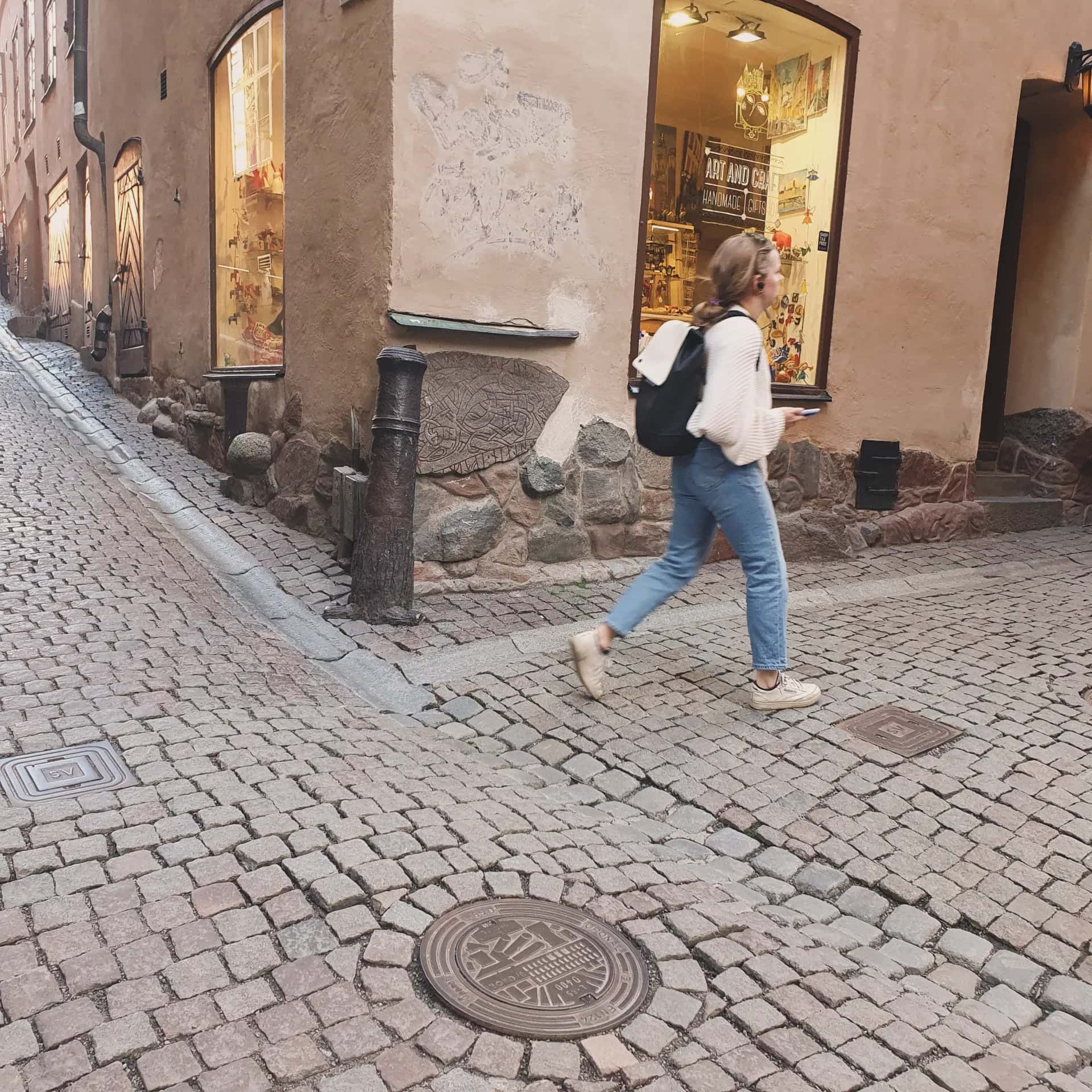 Kvinna med ryggsäck går på kullerstensgata. Butiksfönster med hantverk i gammal byggnad till höger och stigande gata till vänster