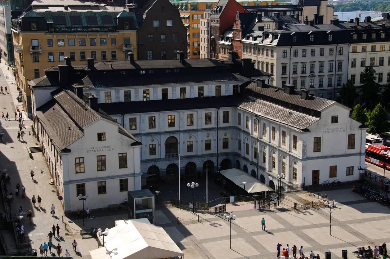 Stockholms Stadsmuseum, en stor vit byggnad med svart tak, sedd från ovan. Människor på torget framför